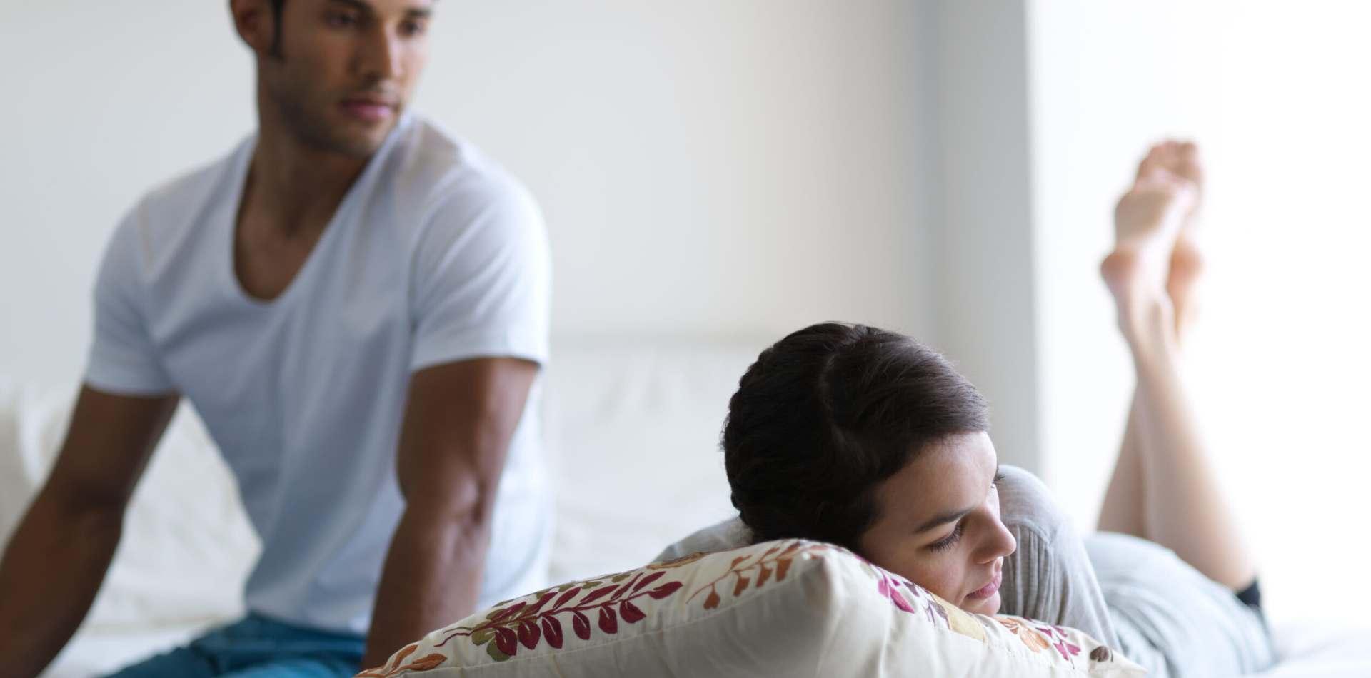 Man sitting on the edge of a bed looking thoughtfully at a woman who is lying on her stomach, resting her head on a pillow, suggesting emotional distance or contemplation.