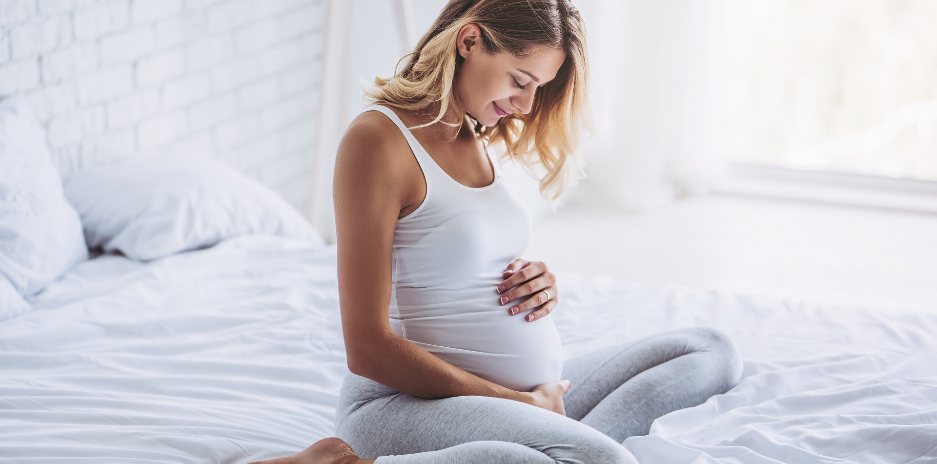 Pregnant woman sitting on a bed, gently cradling her belly and looking at it with a peaceful expression in a bright, airy room.